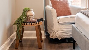 Een gezellige woonkamer met een bank, een klein houten krukje met een plant, een lijstwerk aan de muur en een raam met gordijnen.