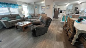 A spacious living room with a grey sofa set, a coffee table, and wicker chairs. A kitchen with white cabinets and a dining area with a table and chairs is visible in the background.
