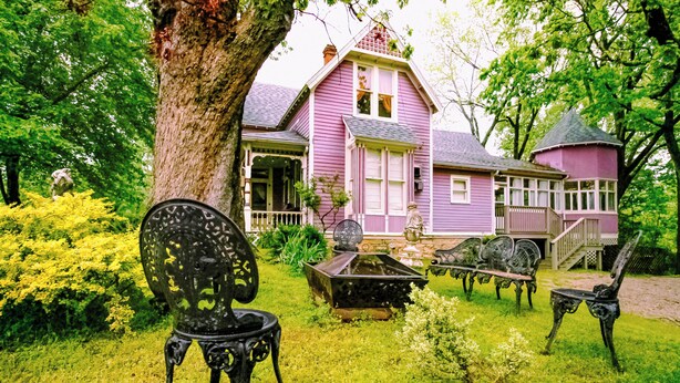 Une maison rose avec un porche, entourée de verdure et agrémentée de mobilier d’extérieur.