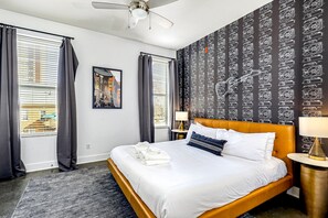 Un cuarto de hotel con una cama grande, un ventilador de techo, una ventana con cortinas, una pared con papel tapiz estampado y un cuadro enmarcado en la pared.