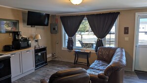 A small living room with a sofa, a television mounted on the wall, a coffee maker, a microwave, and a window with curtains.