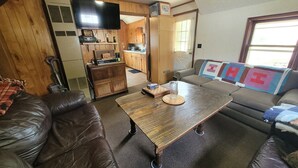 Ein gemütliches Wohnzimmer mit einer braunen Ledersofa, einem Holztisch und einem Sofa mit einer farbenfrohen Stegdecke. Ein Fernseher ist an der Wand befestigt, und im Hintergrund ist eine Küche zu sehen.