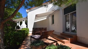 Un patio con una mesa y sillas de madera, rodeado de plantas en macetas y un edificio blanco con puerta de cristal.
