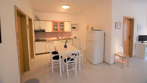 Una cocina compacta con armarios blancos, una mesa de comedor con sillas, un refrigerador y una mesita auxiliar con una lámpara.