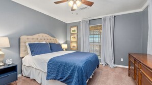 Une chambre à coucher avec un lit doté d’une tête de lit capitonnée, une commode en bois, une table de chevet avec une lampe et un ventilateur de plafond.