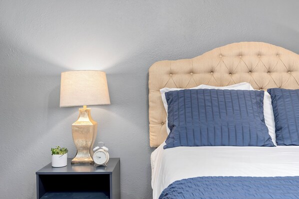 A neatly made bed with a tufted headboard, a bedside table with a lamp, a clock, and a small plant.