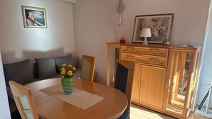 Un comedor con una mesa redonda de madera, sillas y un jarrón con flores amarillas. Se observa un aparador de madera con puertas de cristal y una fotografía enmarcada encima.