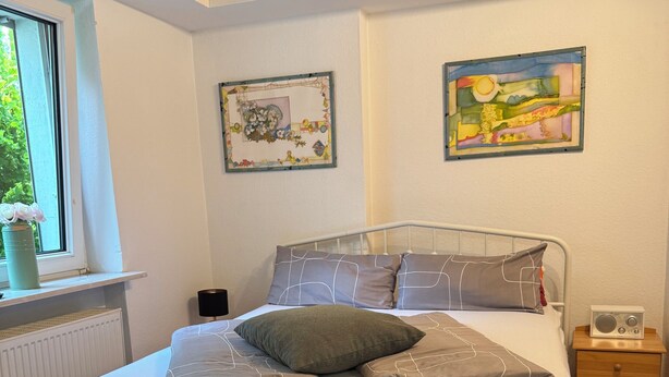 Un dormitorio con una estructura de cama metálica blanca, ropa de cama gris y dos cuadros colgados en la pared.