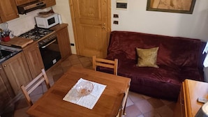Una cocina compacta con armarios de madera, un microondas y una estufa. Hay una mesa de comedor con un bol de vidrio y un mantel de encaje. Un sofá de terciopelo rojo con un cojín dorado está apoyado en la pared.
