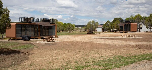 Two modern, brown, glass-enclosed cabins with picnic tables, set in a dry, grassy area with trees and a distant hill.
