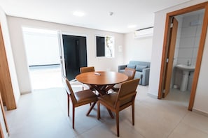 A modern living room with a round wooden table and four chairs, a grey sofa, and a small bathroom visible through an open door. - ROTA DO SOL APART HOTEL (Guaratuba)