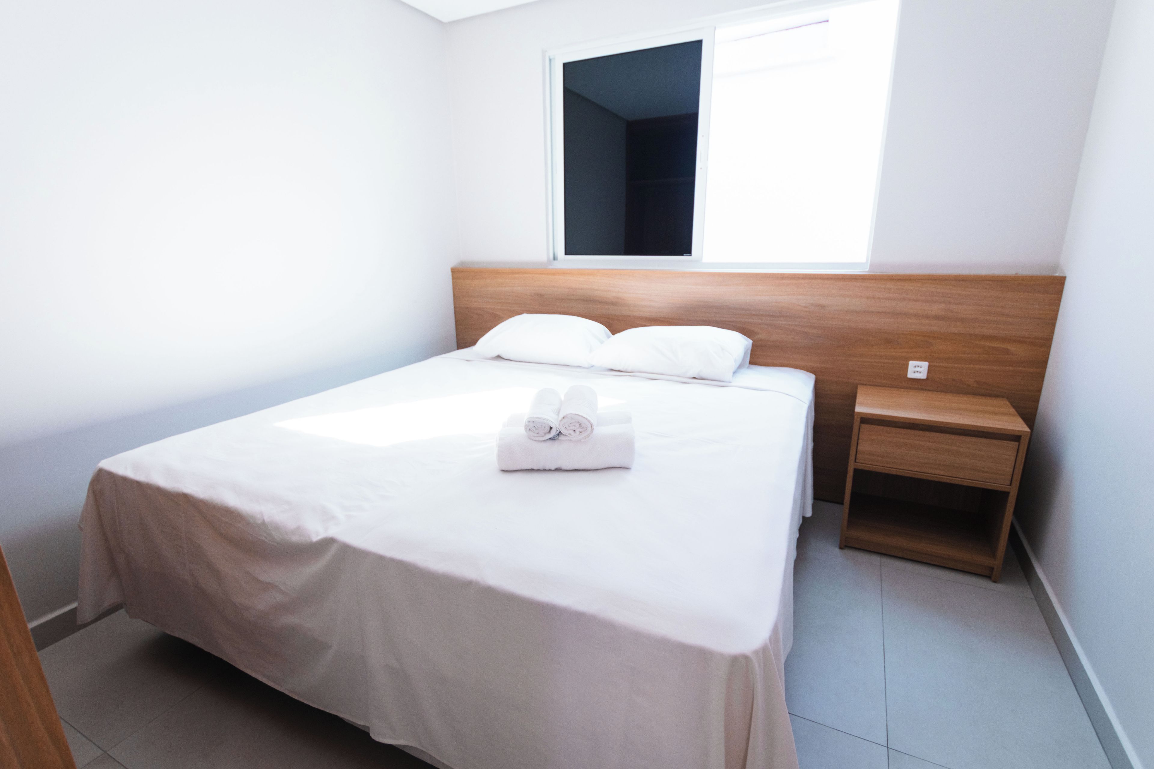 A neatly made bed with white linens and pillows, a wooden headboard, a bedside table with a lamp, and a window with a view of the sky.