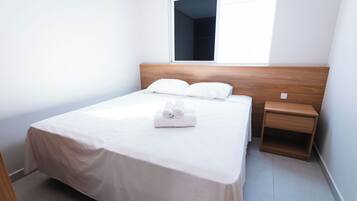 A neatly made bed with white linens and pillows, a wooden headboard, a bedside table with a lamp, and a window with a view of the sky.