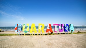 Un letrero en la playa con la palabra 'QUARATUBA' escrita con letras coloridas y varios iconos, frente a un cielo azul despejado y el océano.