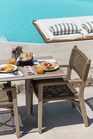 Una mesa de comedor al aire libre con alimentos para el desayuno, una silla de mimbre y una piscina al fondo.