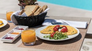 Une table dressée pour le petit-déjeuner avec une assiette d’œufs, de tomates et de légumes verts, un verre de jus d’orange et un panier de pain, le tout placé près d’une piscine.