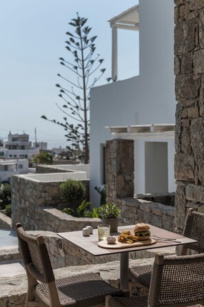 An outdoor dining area with stone walls, wicker chairs, and a table set with a burger, fries, and a drink. - Arocaria Hotel (Mykonos)