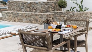 A dining table with wicker chairs, a stone wall, and a view of the sea. - Arocaria Hotel (Mykonos)