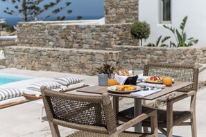 A dining table with wicker chairs, a stone wall, and a view of the sea. - Arocaria Hotel (Mykonos)