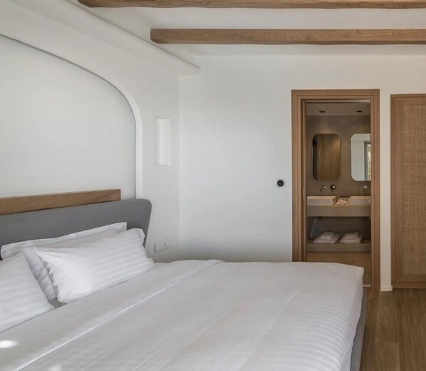 A bedroom with a bed, a wooden headboard, a built-in wardrobe, and a bathroom with a mirror and sink. - Arocaria Hotel (Mykonos)