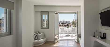 A modern bedroom with a large bed, a seating area with a bench, a TV mounted on the wall, and a balcony with a table and chairs.