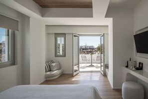A modern bedroom with a large bed, a seating area with a bench, a TV mounted on the wall, and a balcony with a table and chairs. - Arocaria Hotel (Mykonos)