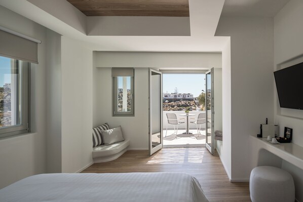 Un dormitorio moderno con una cama grande, una zona de estar con un banco, un televisor montado en la pared y un balcón con mesa y sillas.