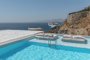 A pool with lounge chairs and a view of the sea and a cruise ship. - Arocaria Hotel (Mykonos)