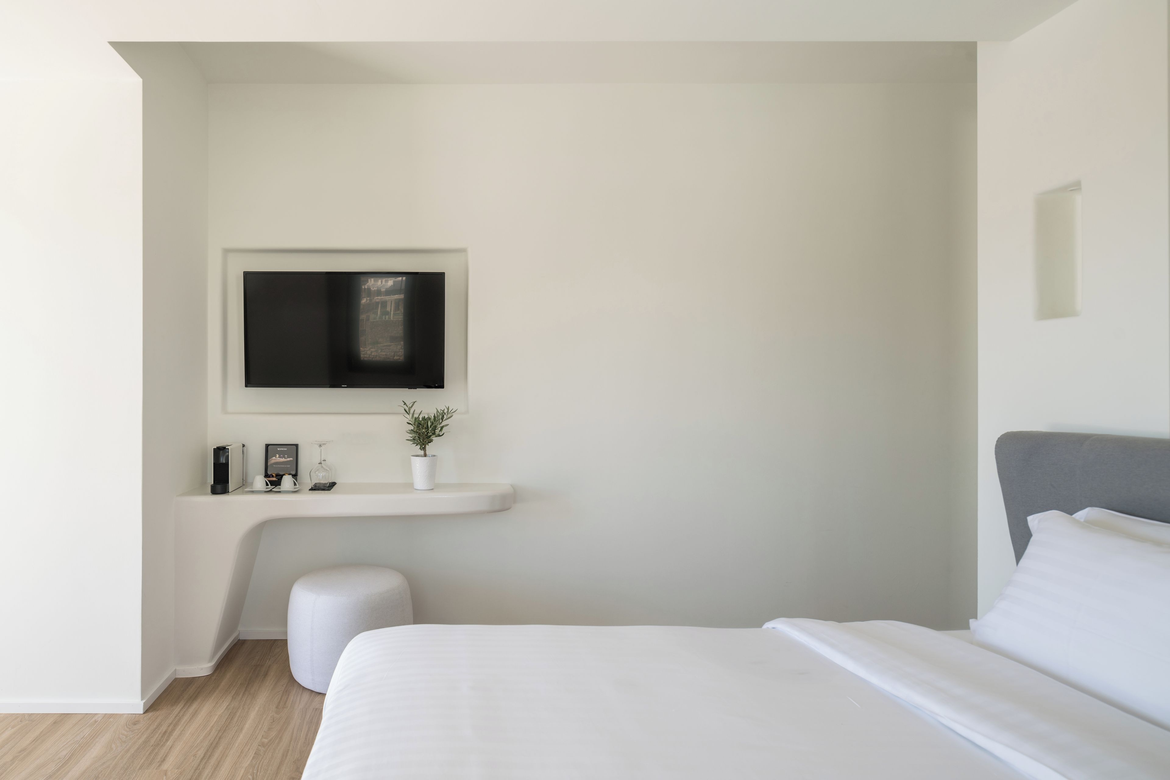 A modern bedroom with a bed, a TV mounted on a white wall, a small white bench, and a potted plant.