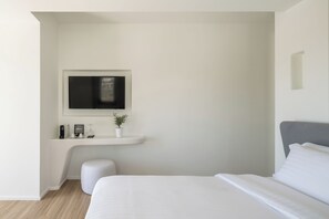 A modern bedroom with a bed, a TV mounted on a white wall, a small white bench, and a potted plant. - Arocaria Hotel (Mykonos)