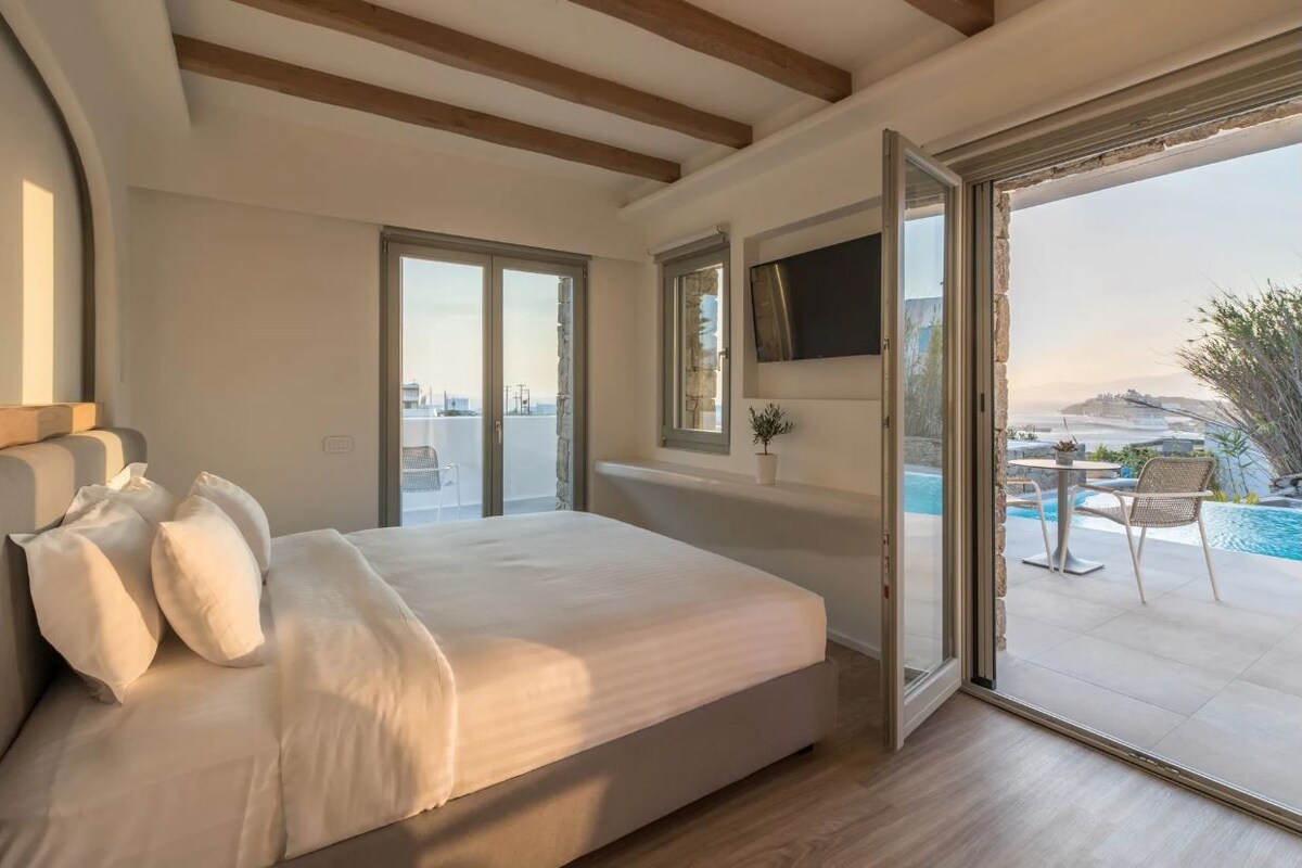 A bedroom with a large bed, a TV mounted on the wall, and a sliding glass door leading to a balcony with a pool.