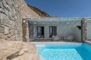 A stone-walled pool area with a clear blue pool, stone pillars, and a covered seating area. - Arocaria Hotel (Mykonos)