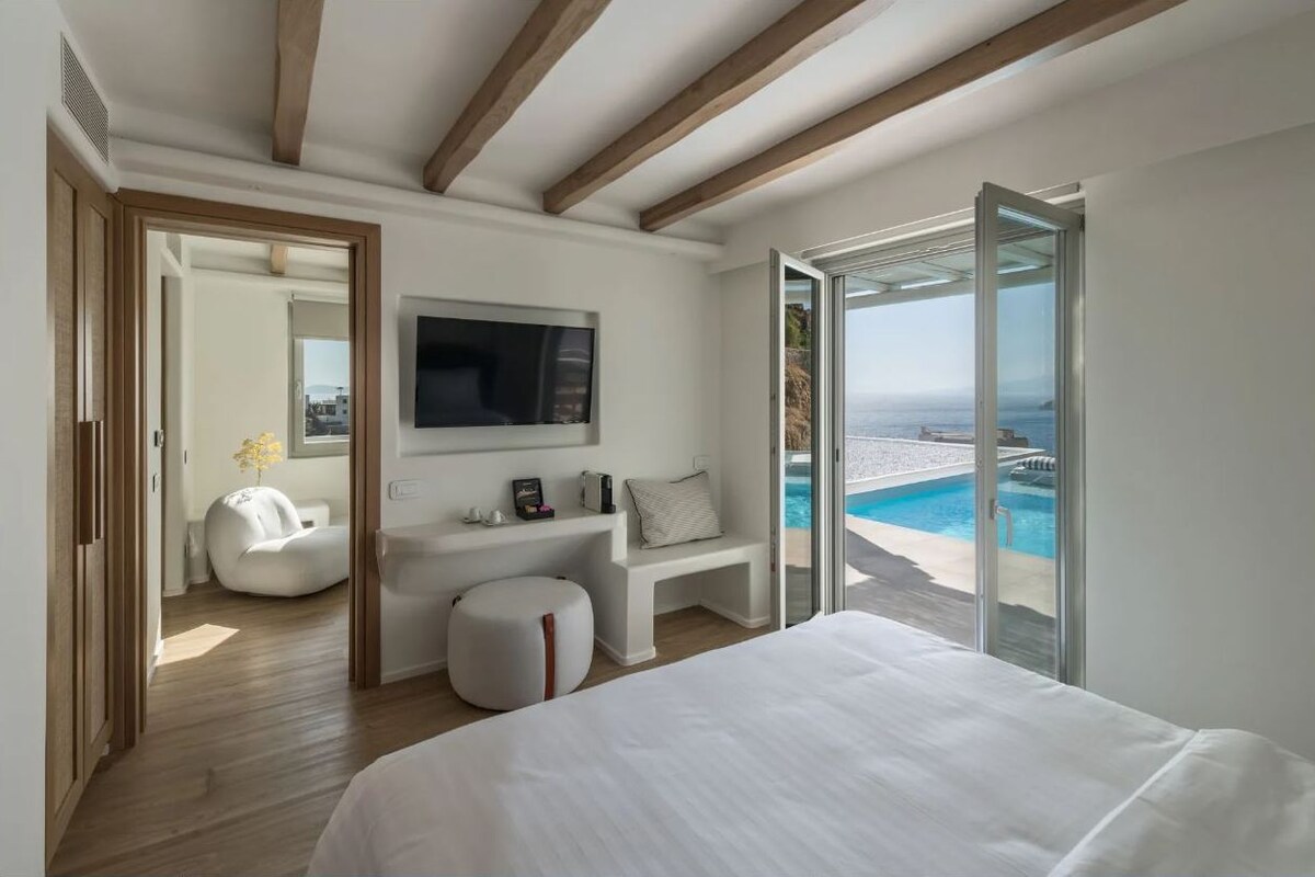 A modern bedroom with a large bed, a TV, and a view of a pool and ocean.