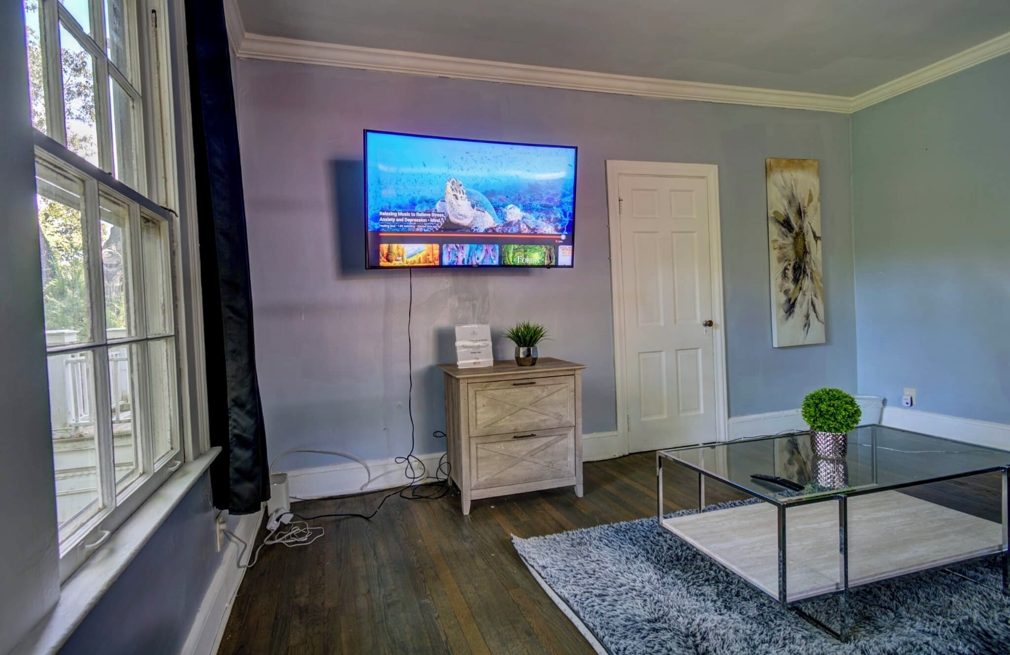 Uma sala de estar com televisão de tela plana na parede, um armário de madeira, uma mesa de centro de vidro e um tapete azul.