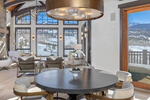 A modern interior with a round dining table, comfortable seating, large windows with a snowy mountain view, and a high ceiling with exposed beams.