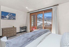 A bedroom with a bed, a dresser, a chair, and a view of mountains through a large window.