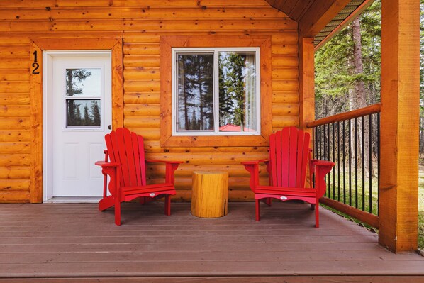 Una cabaña de troncos con una terraza de madera, dos sillones Adirondack rojos, una mesita de madera y una puerta blanca con el número 12.