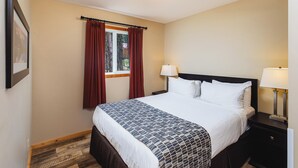 A hotel room with a bed, two bedside lamps, a window with curtains, a framed picture on the wall, and a wooden floor. - Single Bedroom Unit in the Mountain (Jasper east)