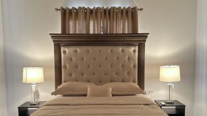 Una cama bien hecha con cabecera capitonada, dos lámparas de noche y una ventana con cortinas.