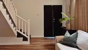 Un salón con una escalera, una puerta oscura, una planta en maceta y un sofá color beige.