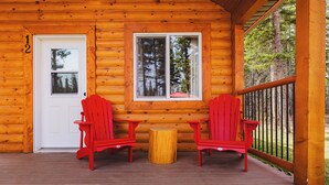 A log cabin with a wooden deck, two red Adirondack chairs, a small wooden table, and a white door with the number 12.
