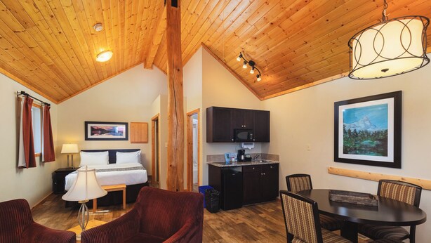 A cozy room with a bed, a desk, a chair, a dining table with chairs, a kitchen area, and a wooden ceiling.