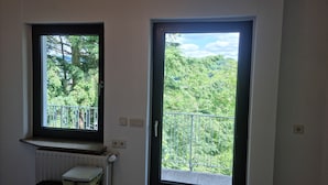 Une pièce dotée de deux fenêtres : l’une offre une vue sur les arbres et un balcon, et l’autre donne sur un feuillage dense.