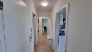 Un couloir étroit aux murs blancs et au plancher en bois, qui mène à une pièce dotée d’un coin cuisine.