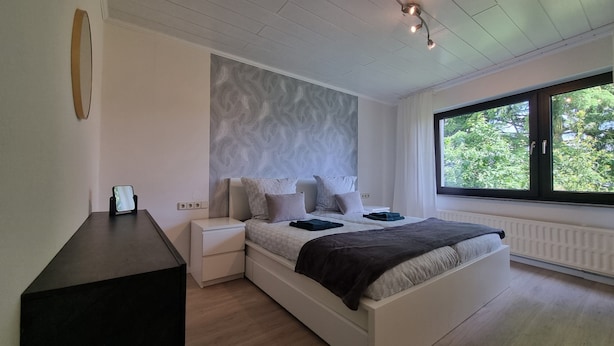 Une chambre à coucher comprenant un lit, des tables de chevet, une commode, une fenêtre donnant sur un espace verdoyant et un luminaire au plafond.