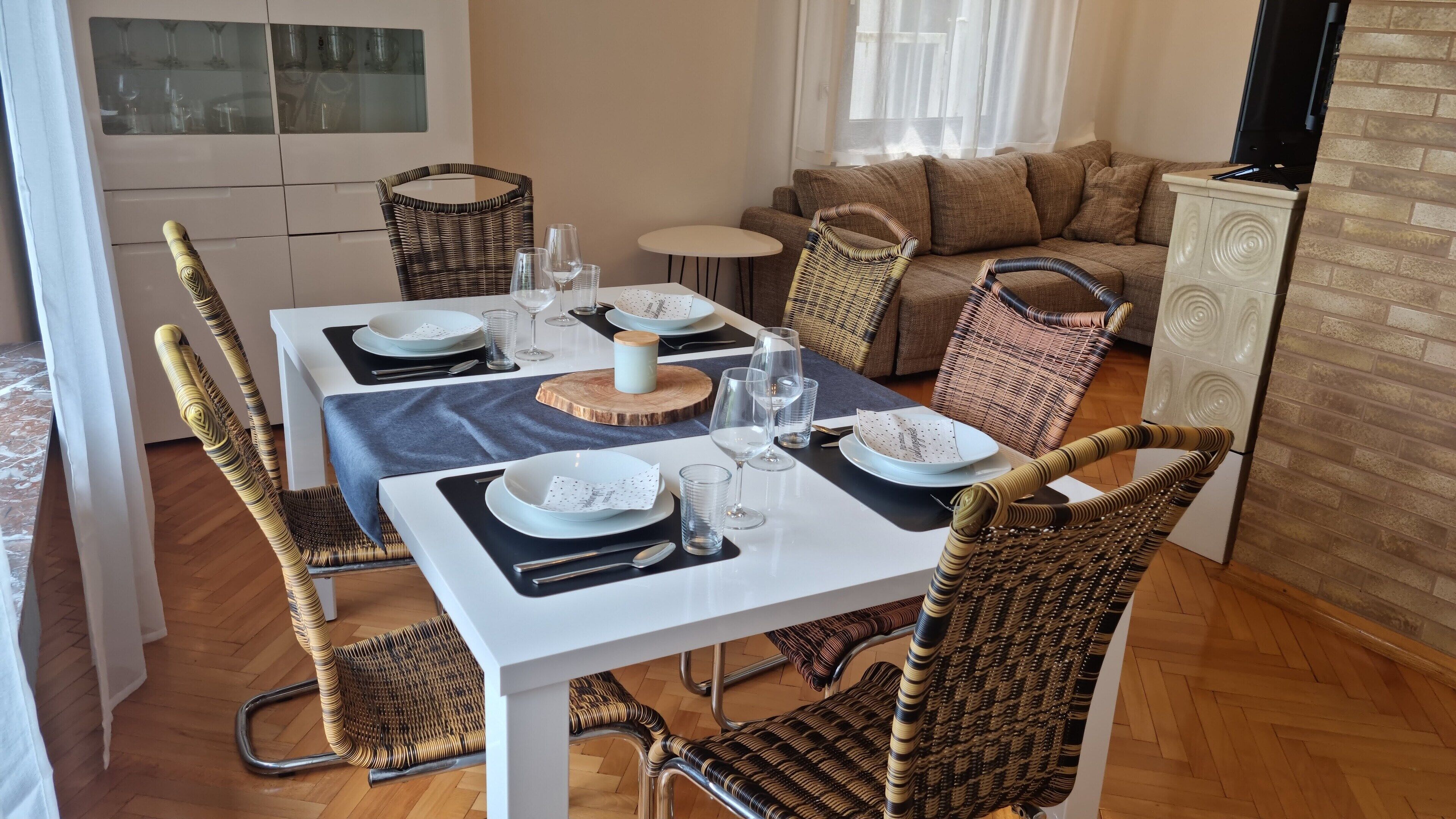 A dining area with a white table set for four, surrounded by wicker chairs, a sofa, and a television.