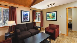 Un salón acogedor con un sofá marrón oscuro, un sillón rojo y una mesa de centro de madera. Hay dos ventanas con cortinas rojas y obras de arte en las paredes.