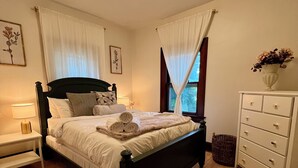 En pænt redt seng med puder og et sammenrullet håndklæde, et sengebord med en lampe, en kommode og et vindue med gardiner.