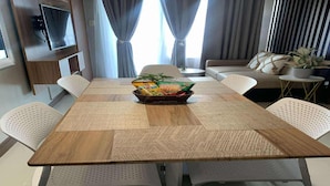 Un salón moderno con una mesa de comedor de madera, sillas blancas perforadas, un sofá, un televisor montado en la pared y una planta sobre una mesita auxiliar.