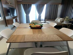 Un salon moderne avec une table à manger en bois, des chaises blanches perforées, un canapé, un téléviseur fixé au mur et une plante posée sur une table d’appoint.